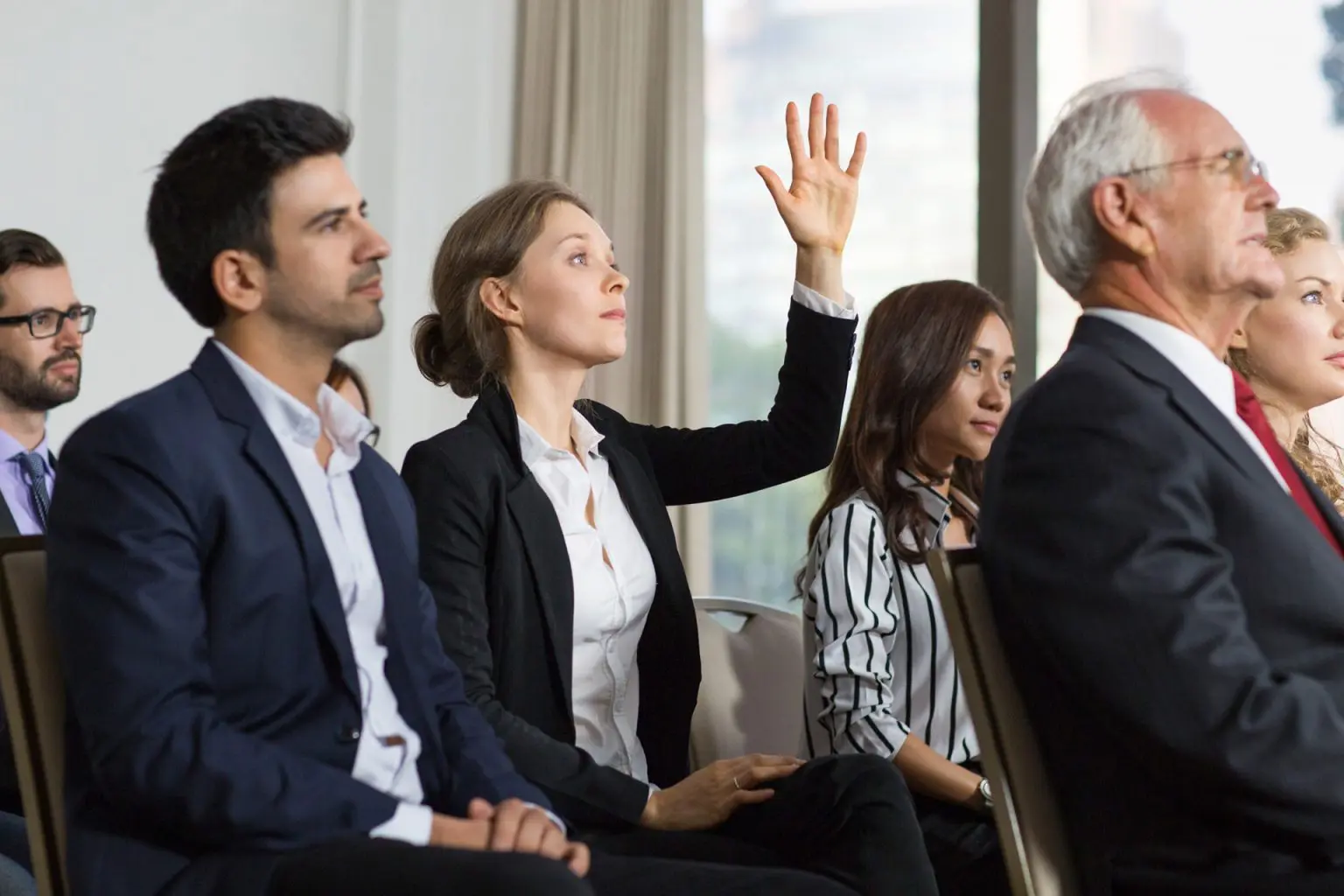 A woman raises her hand during a formal meeting while others listen attentively.