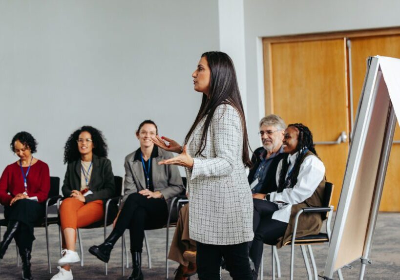 A woman presenting to an attentive audience in a professional setting.
