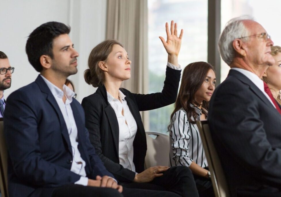 A woman raises her hand during a formal meeting while others listen attentively.